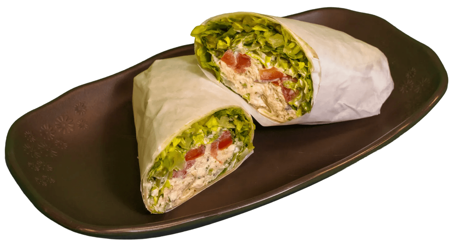 Two halves of a chicken or tuna salad wrap, cut diagonally, filled with crisp green lettuce, diced red tomatoes, and creamy salad, partially wrapped in white paper, resting on a dark brown, subtly patterned plate.