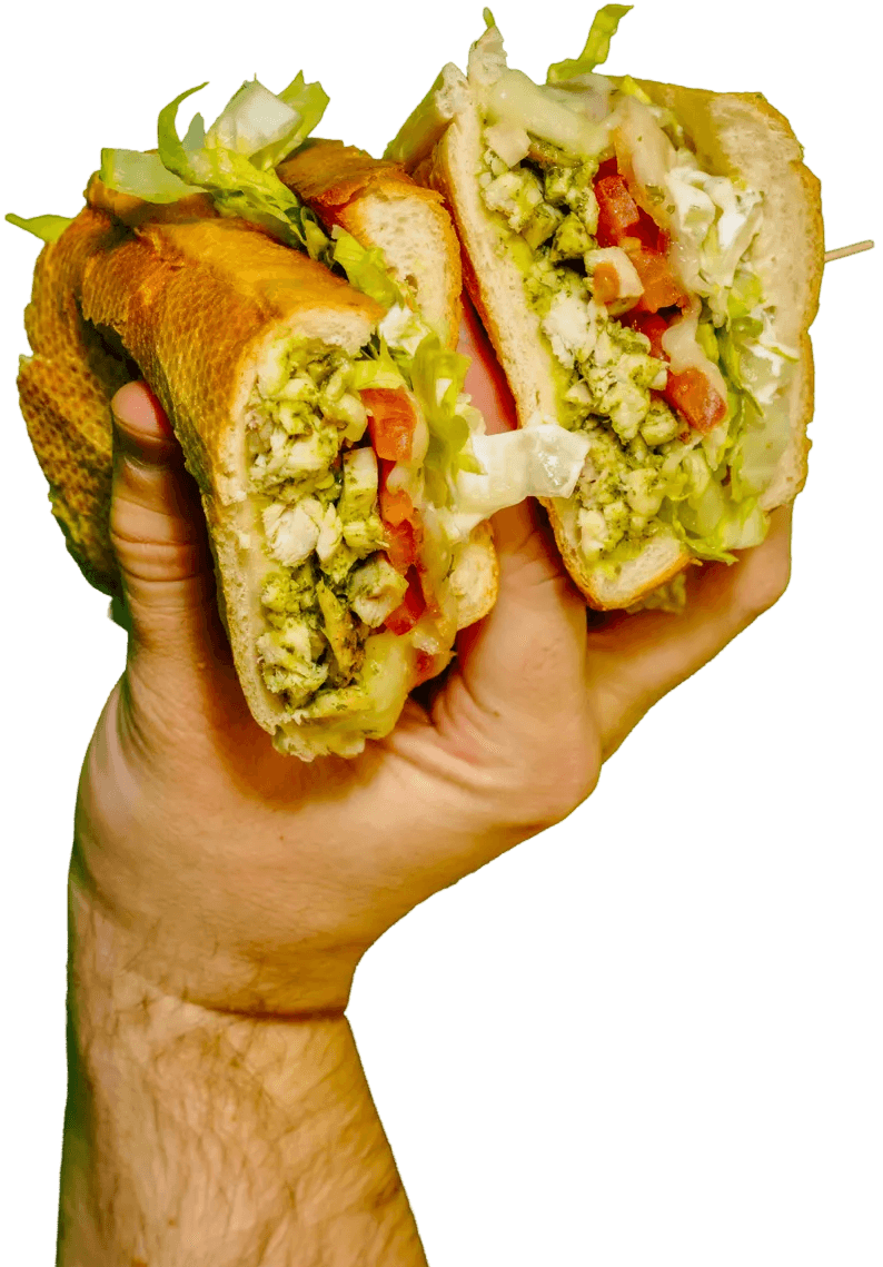 A hand holds a chicken pesto sandwich, cut in half, revealing chicken chunks, green pesto, lettuce, tomatoes, and melted cheese on a baguette.